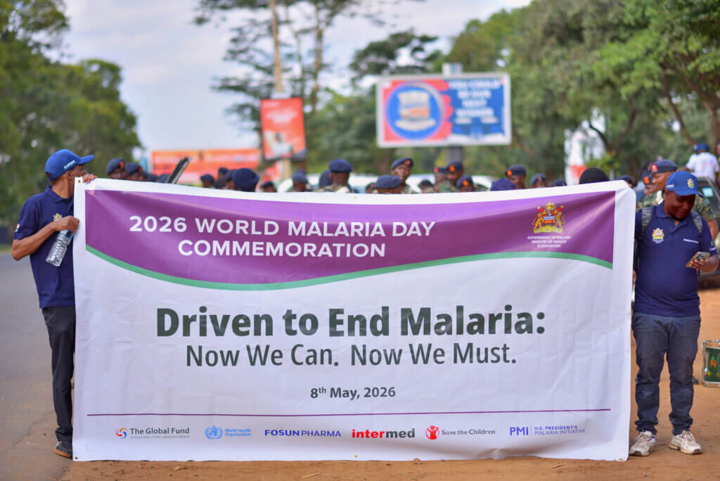 Participants carry a large banner during the 2026 World Malaria Day Commemoration march. The banner features the campaign message “Driven to End Malaria: Now We Can. Now We Must.” alongside logos from the Malawi Ministry of Health and Sanitation, WHO, The Global Fund, Save the Children, PMI, Fosun Pharma, and other partners. Uniformed personnel and attendees gather in the background as the procession moves through a public roadway.