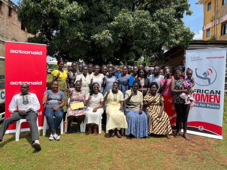 Group photo of approximately 40 women and men seated and standing outdoors under a large tree, with ActionAid and African Women Voices for Peace banners displayed on either side