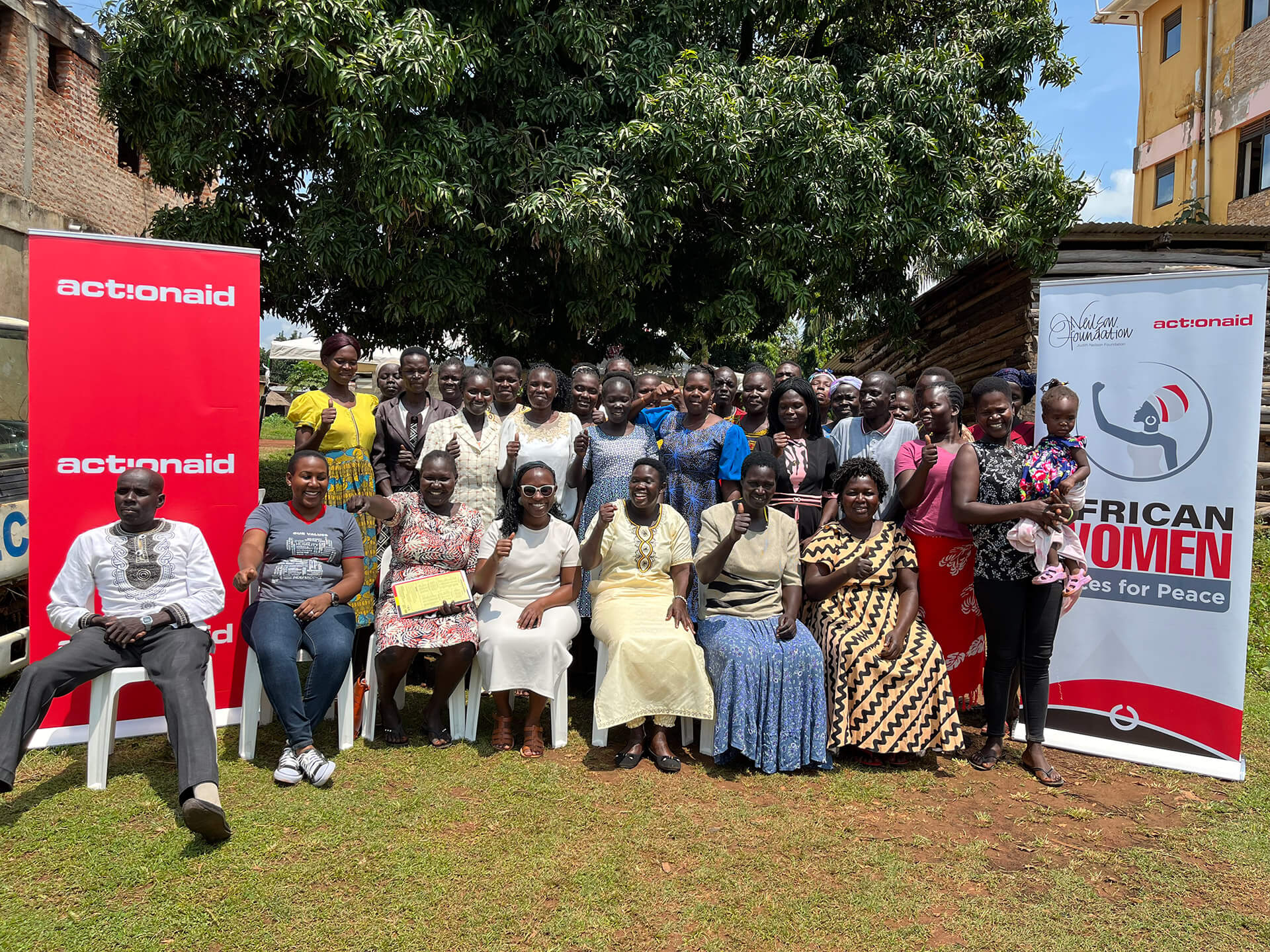 Group photo of approximately 40 women and men seated and standing outdoors under a large tree, with ActionAid and African Women Voices for Peace banners displayed on either side