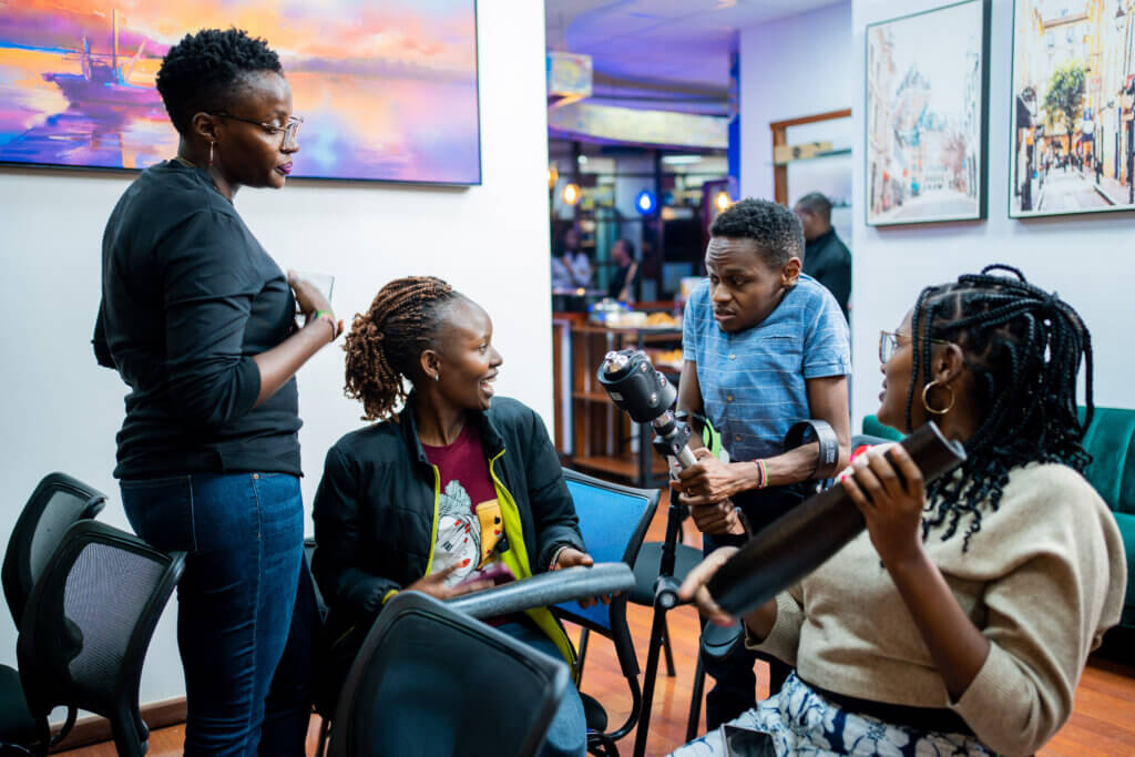 our people gather indoors in a casual workspace or studio, talking and laughing while handling equipment. One person holds a camera mounted on a handheld stabilizer, while another holds a long cylindrical object, possibly part of filming or audio gear. The others watch and engage in conversation, smiling. The room features chairs, framed artwork on the walls, and a softly lit background with additional people and activity, suggesting a collaborative or creative setting.