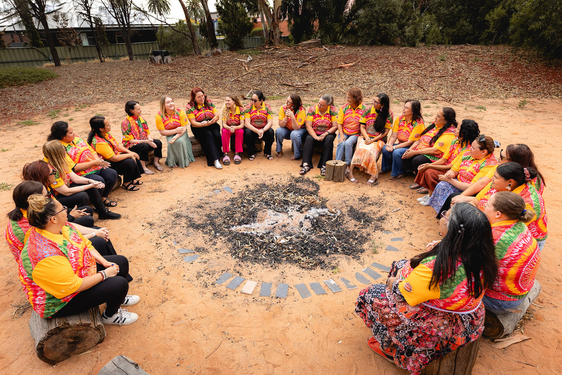 A group of people sit in a circle outdoors on logs arranged around a central fire pit, engaged in conversation. They wear matching brightly patterned shirts, suggesting they are part of the same group or event. The setting is a natural, sandy area with trees and vegetation nearby, creating a calm, communal atmosphere focused on discussion and connection.