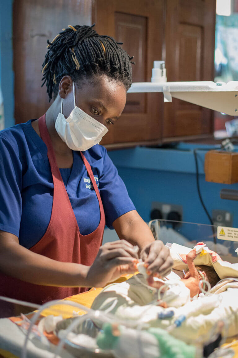A healthcare worker wearing blue scrubs, a protective apron, and a face mask carefully tends to a newborn infant under bright medical lighting. The worker’s hands are focused on adjusting tubing or monitoring equipment attached to the baby. The setting appears to be a neonatal care unit, with clinical equipment and a protective enclosure visible around the infant, conveying a scene of attentive medical care.
