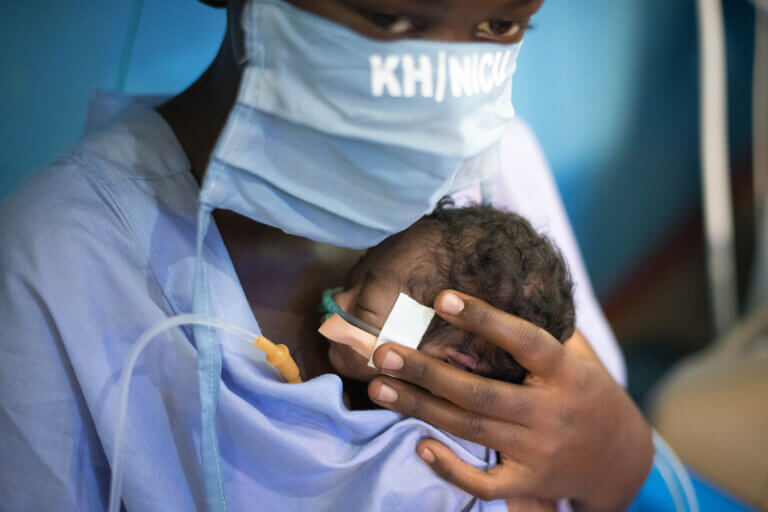 A healthcare worker wearing a face mask and light blue scrubs holds a very small newborn infant gently against their chest in a skin-to-skin care position. The baby has a nasal tube secured to the face and rests closely against the caregiver’s body while being supported by one hand. The close framing emphasizes careful, intimate medical care in what appears to be a neonatal hospital setting.