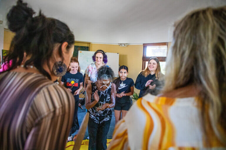 A group of people participate in a lively indoor movement or dance session, standing in a circle and clapping while stepping in rhythm. Some are barefoot, and the atmosphere feels informal and collaborative. The room includes a whiteboard, tables, and a wall-mounted air conditioner, suggesting a workshop or training space. Participants appear engaged and smiling as they move together, creating a sense of shared energy and community.