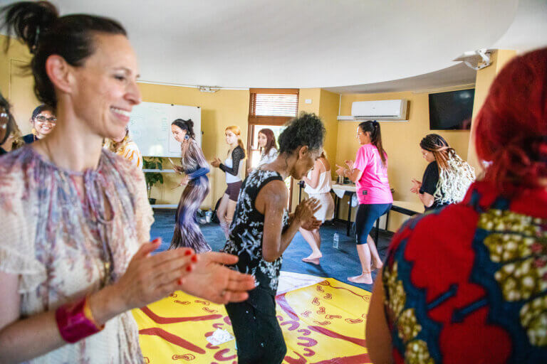 A group of people participate in a lively indoor movement or dance session, standing in a circle and clapping while stepping in rhythm. Some are barefoot, and the atmosphere feels informal and collaborative. The room includes a whiteboard, tables, and a wall-mounted air conditioner, suggesting a workshop or training space. Participants appear engaged and smiling as they move together, creating a sense of shared energy and community.