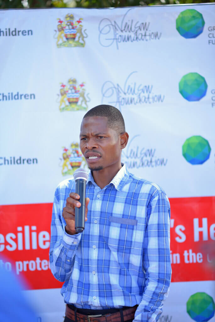 A man in a blue checkered shirt speaks into a microphone at an outdoor event, standing in front of a branded backdrop featuring the Malawi government crest, Save the Children, the Green Climate Fund, and the Nilson Foundation. The scene appears to be part of a public awareness or community engagement event related to climate resilience or health initiatives.