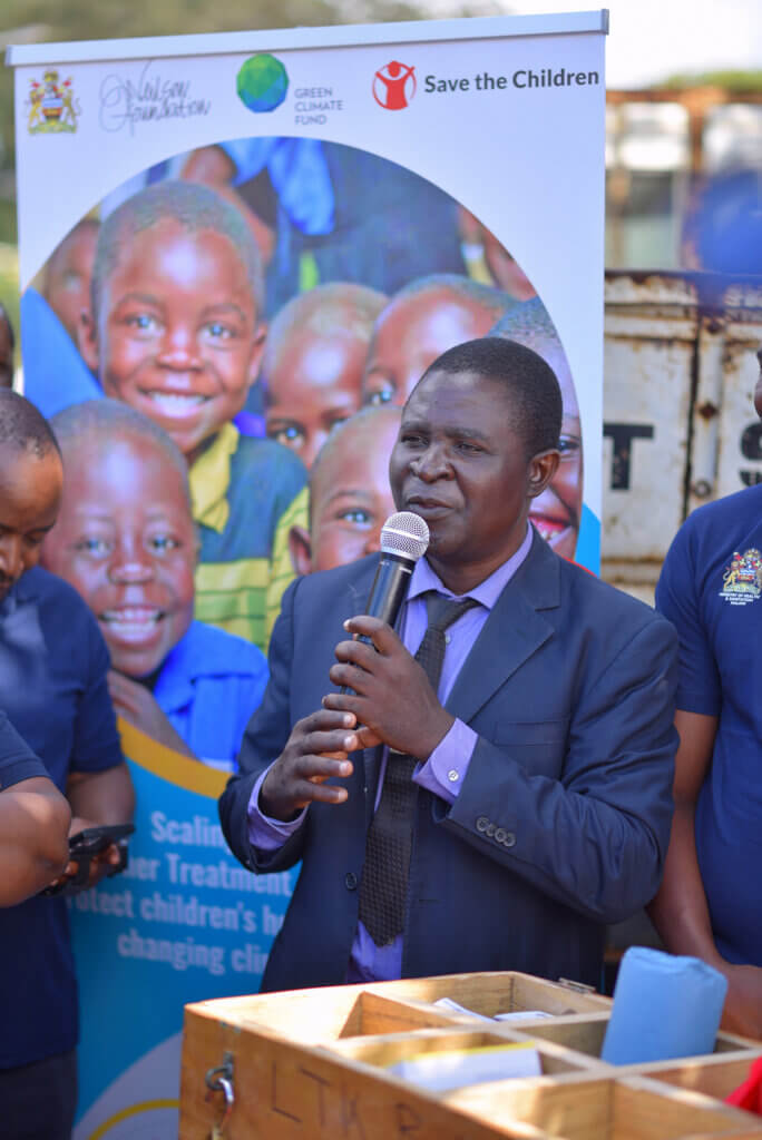 A man in a dark suit and tie speaks into a microphone beside a large Save the Children and Green Climate Fund display featuring smiling children. Other attendees stand nearby, and the image captures a formal speech during a community or health-focused event.
