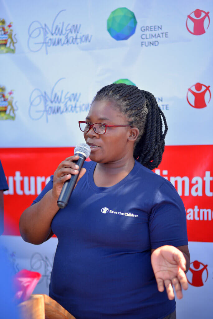 A woman wearing glasses and a Save the Children shirt speaks into a microphone at an outdoor event. She stands in front of a branded backdrop featuring the Nilson Foundation and Green Climate Fund logos, appearing to address attendees during a public health or climate resilience campaign gathering.