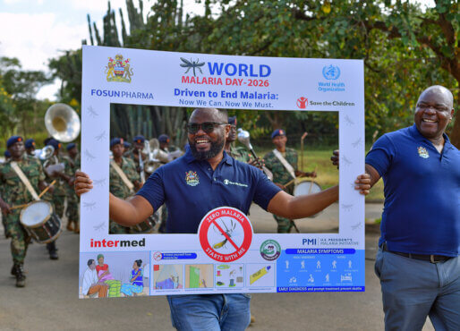 A person walks outdoors holding a large promotional frame for “World Malaria Day 2026,” smiling toward the camera while framed within the sign. The display includes logos from organizations such as the World Health Organization and Save the Children, along with malaria prevention messaging and illustrations. Another person walks beside them, also smiling, while a marching band in uniform plays instruments in the background. The scene appears to be part of a public awareness parade or community event focused on malaria prevention.