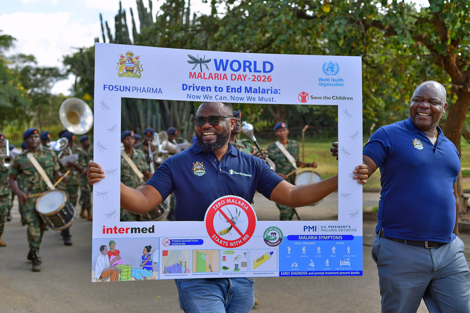 A person walks outdoors holding a large promotional frame for “World Malaria Day 2026,” smiling toward the camera while framed within the sign. The display includes logos from organizations such as the World Health Organization and Save the Children, along with malaria prevention messaging and illustrations. Another person walks beside them, also smiling, while a marching band in uniform plays instruments in the background. The scene appears to be part of a public awareness parade or community event focused on malaria prevention.