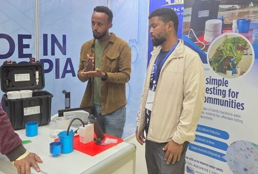 Two representatives demonstrate a portable water testing and purification system at an exhibition booth. Surrounded by sample containers, equipment cases, and informational displays, the presenters engage attendees while showcasing practical water quality solutions designed for community use and accessible field testing.