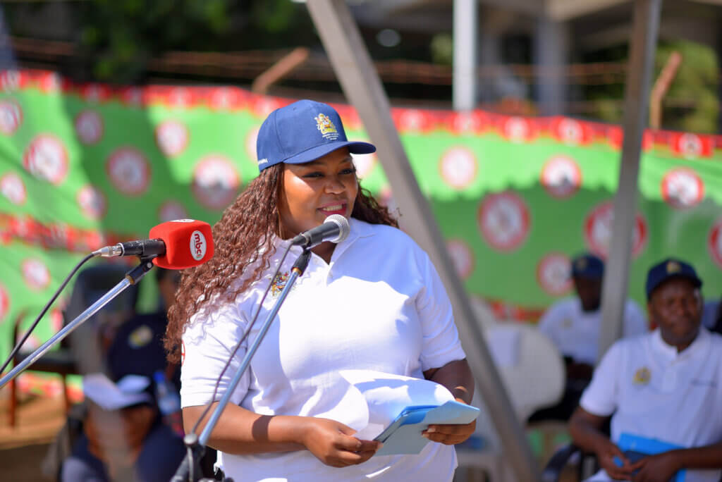 A woman wearing a white polo shirt and navy cap addresses a crowd from a microphone stand while holding notes. Event staff and attendees sit in the background beneath a green malaria-awareness banner, suggesting a formal public health or advocacy gathering.