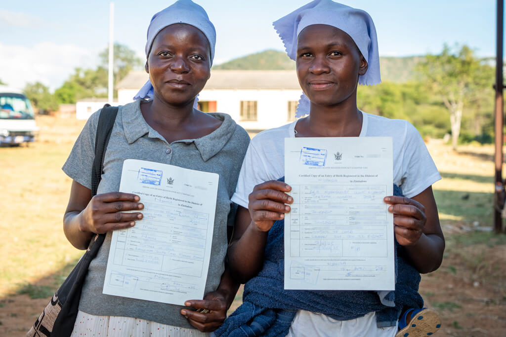 Two women stand outdoors holding certified copies of their birth registration documents, smiling as they present the official certificates to the camera. The image highlights improved access to civil registration services and legal identity documentation, reflecting efforts to strengthen birth registration and ensure individuals can access essential rights and public services.