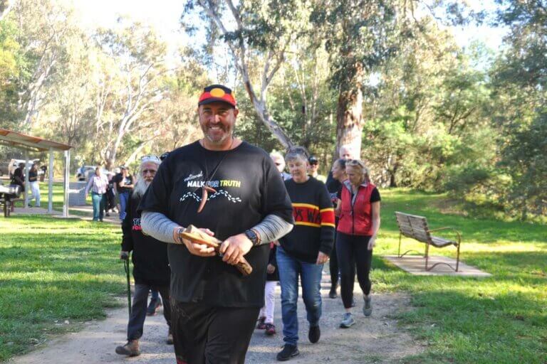 A group of people walk together through a park during a community Walk for Truth event. The lead participant wears a "Walk for Truth" T-shirt and Aboriginal flag cap while carrying clapsticks, with other walkers following along a tree-lined path in a show of solidarity and reconciliation.