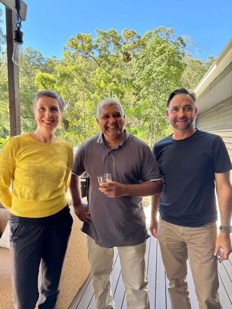 Three people stand smiling together on an outdoor deck surrounded by bushland. They face the camera in a relaxed setting, suggesting a collaborative meeting or informal gathering in a natural environment.