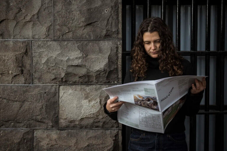 A woman with curly hair stands against a dark stone wall reading an open newspaper titled About Time.