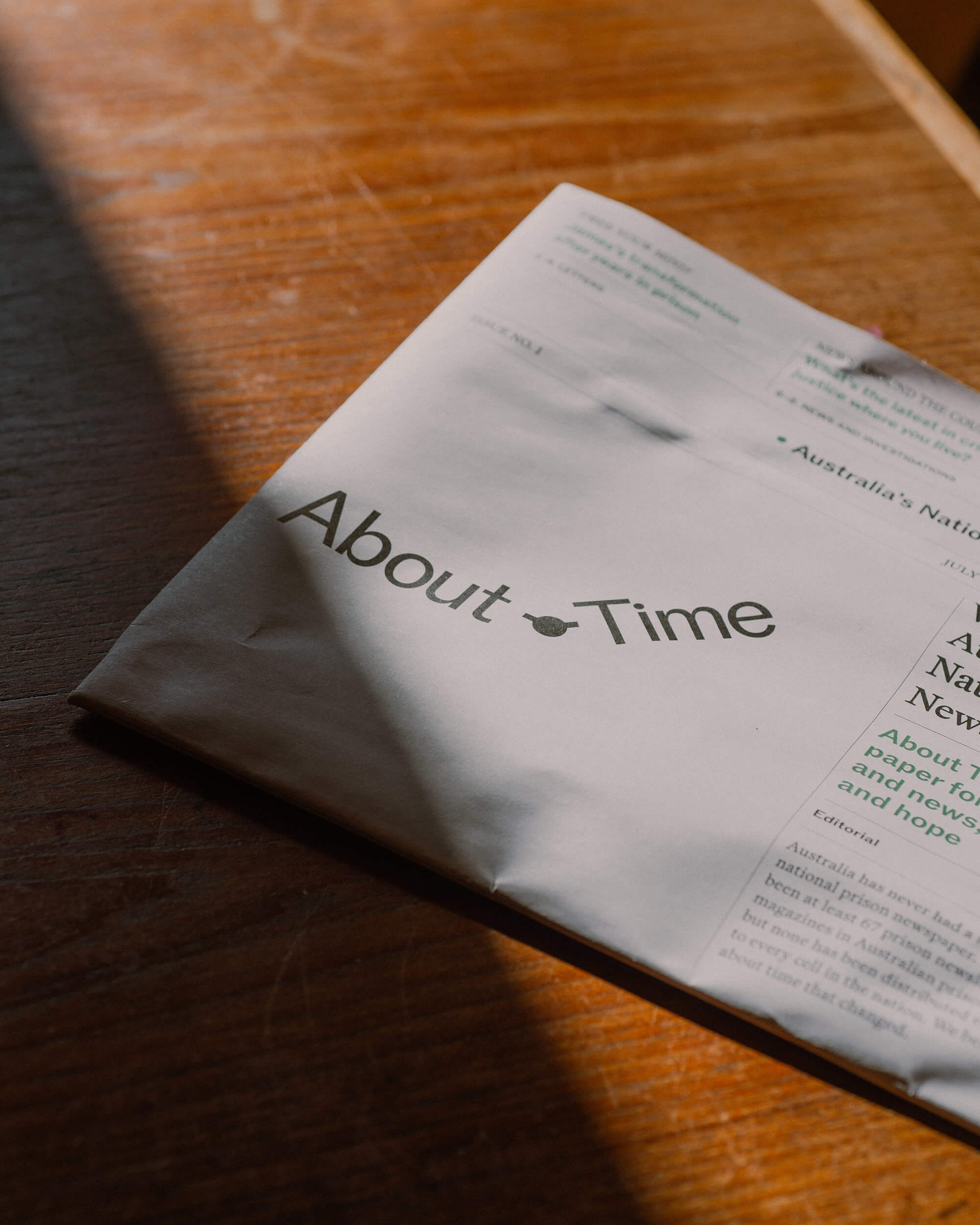 A folded copy of the About Time newspaper rests on a wooden table, illuminated by warm sunlight and shadows.