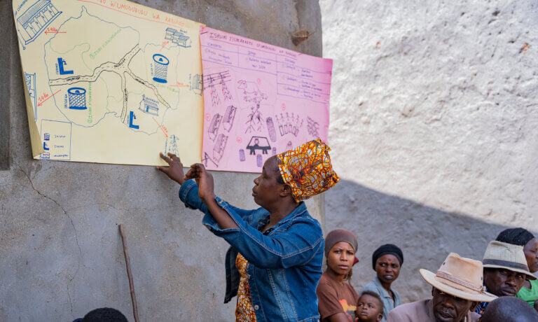 A woman in a patterned headwrap points to hand-drawn community maps and diagrams displayed on a wall while a group of people sit and listen.