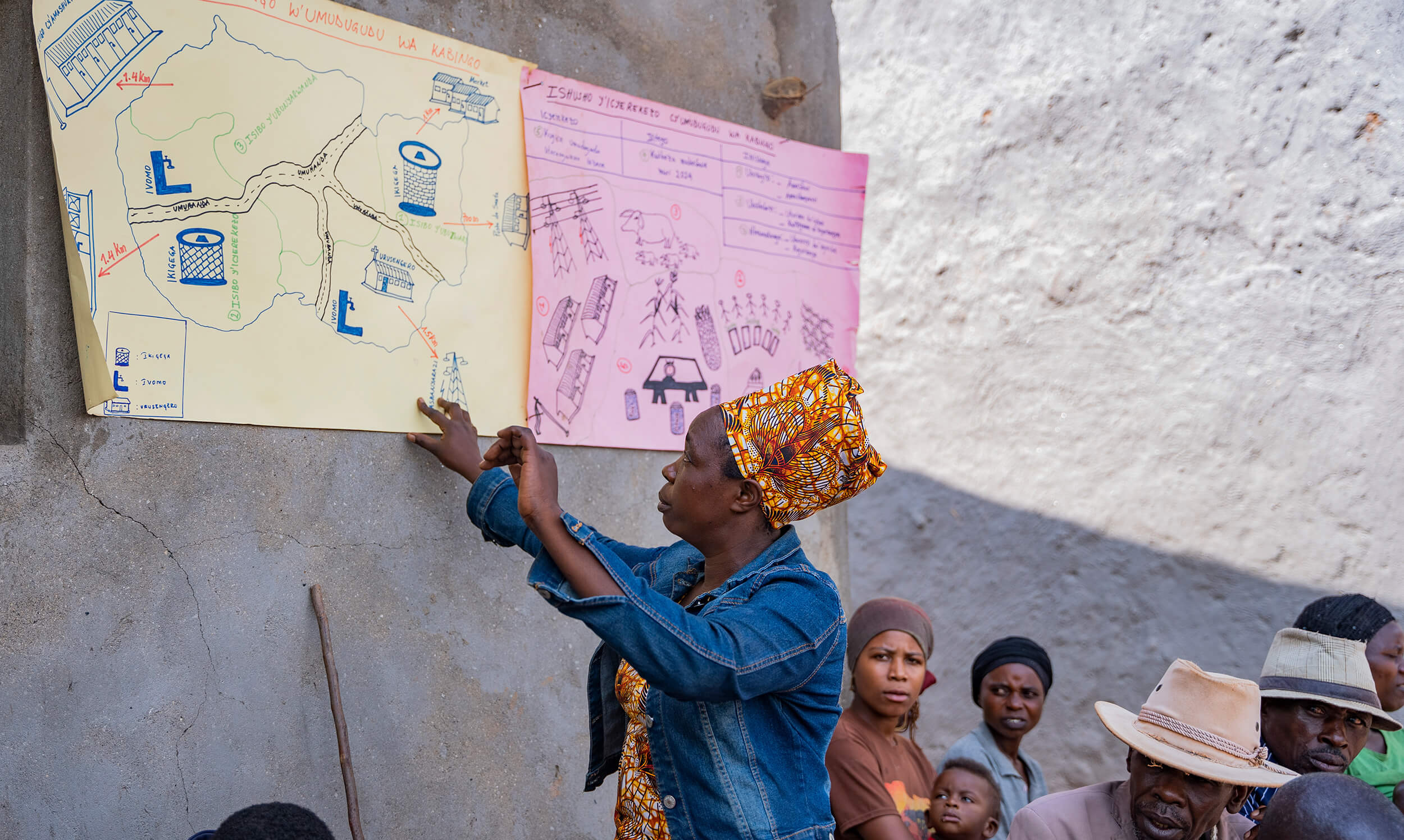 A woman in a patterned headwrap points to hand-drawn community maps and diagrams displayed on a wall while a group of people sit and listen.