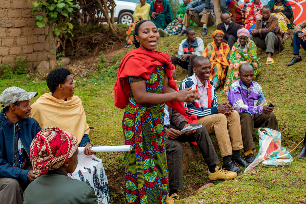 A woman addresses a community gathering outdoors, surrounded by people seated together on a grassy hillside.