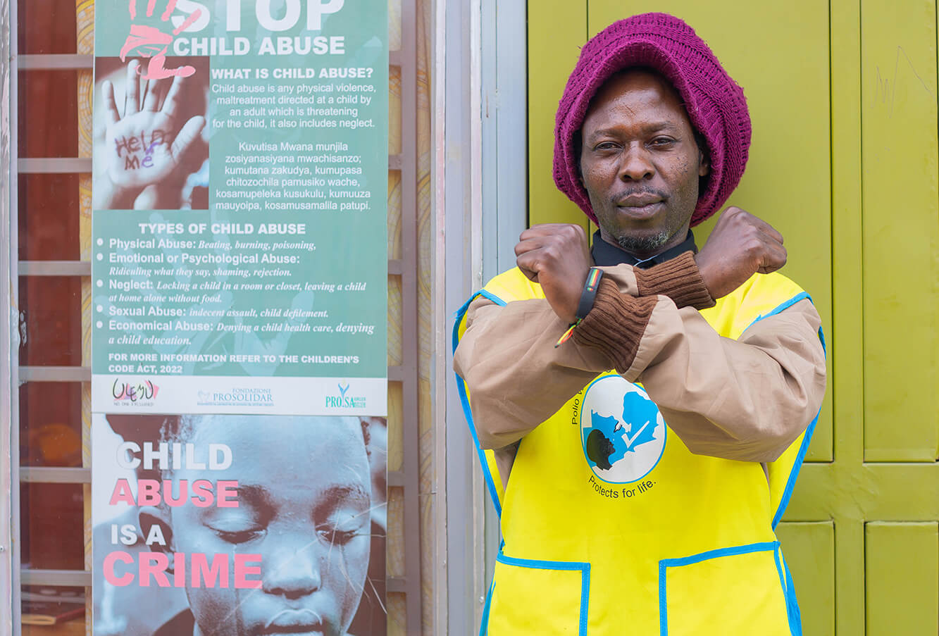 Community health worker stands beside child abuse awareness posters, crossing his arms in a protective gesture.
