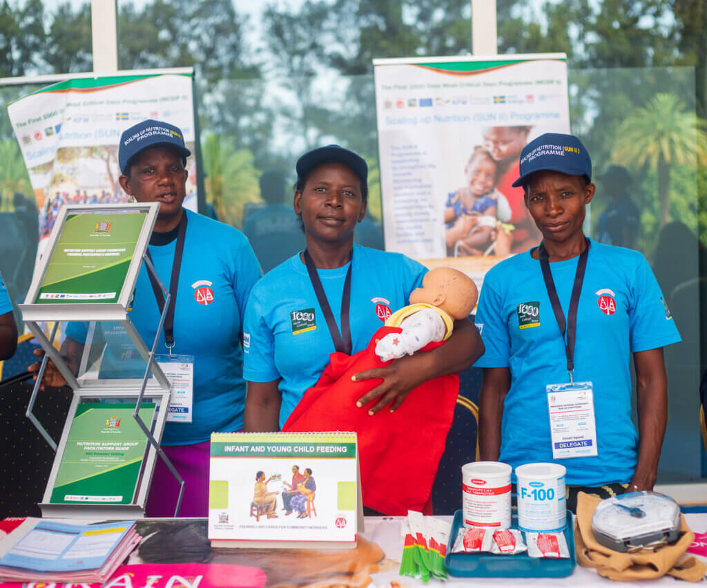 Three community health workers stand at a nutrition information display, with infant feeding resources and training materials.